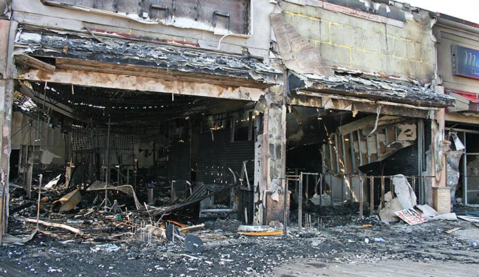 Fire-damaged commercial building with a collapsed roof, charred walls and debris scattered across the entrance