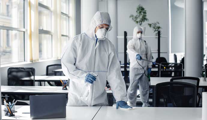 Two experts in protective suits disinfecting office desks and surfaces using spray bottles and cleaning equipment